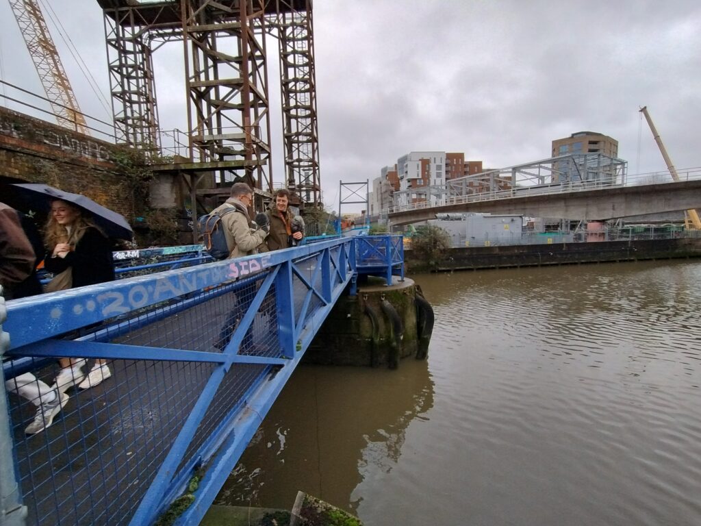 Photo of poet James Wilkes & audio magician Michael Umney plumbing the creek with a hydrophone for the Sounding Deptford project. They stand on a girdered footbridge overlooking rippled waters.