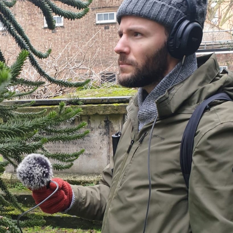 Photo of a trepidatious Joseph Rizzo Naudi holding a furry microphone up to some fir tree branches in the churchyard in Deptford. Joseph wears a grey woolly hat, headphones,  red gloves, a green jacket and a serious expression.